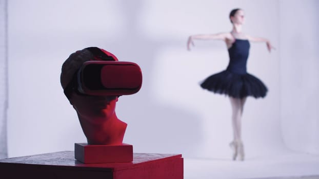 Young woman ballerina in black dress training - a head of greek sculpture in VR glasses on the foreground - red lighting. Mid shot