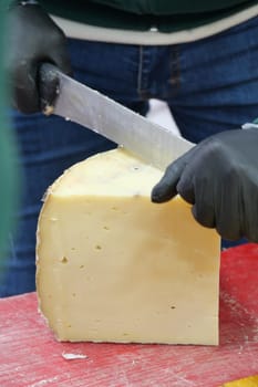A person is skillfully cutting a large block of cheese at a market.