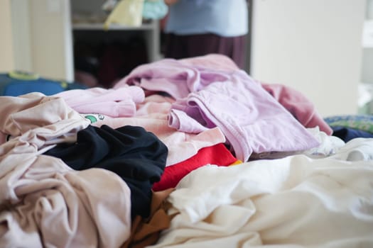 A cluttered pile of clothes is on a bed, with a person organizing nearby.