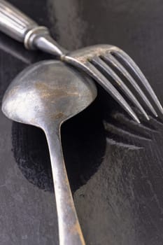 Ancient worn vintage cutlery on the dark background. Crossed black metal spoon and fork in dark tone, Highlighting its texture and design, use it as your Wallpaper, Poster and Copy space Selective focus.