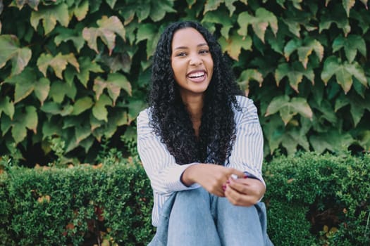 A joyful young woman with delightful curly hair is sitting comfortably in front of vibrant, lush, and green vines, radiating happiness and confidence while enjoying a casual and relaxed setting