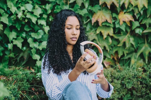 A young woman is peacefully examining and enjoying her stylish headphones, all while being surrounded by vibrant and lush greenery, which beautifully embodies a sense of tranquility and modern style