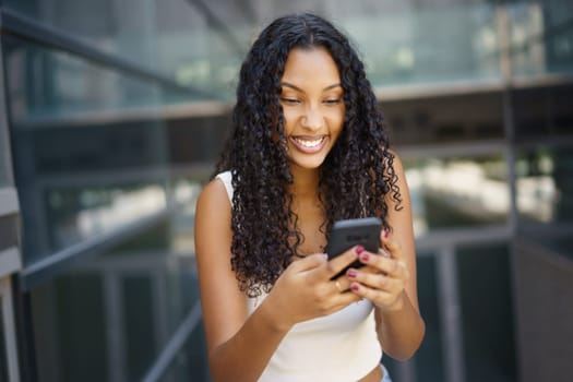 A cheerful young woman with curly hair is outdoors, happily enjoying using her smartphone as she connects with others, showcasing her happiness and a sense of vibrant connection with the world