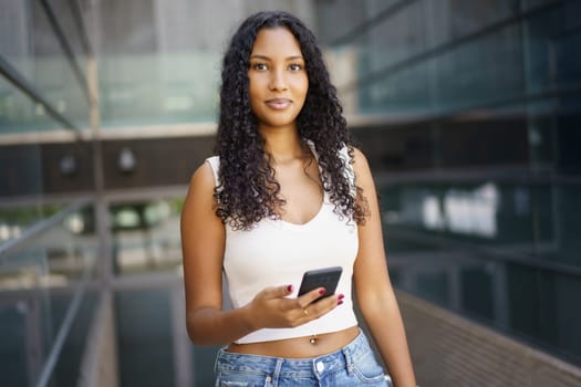 A trendy and stylish young woman dressed in a casual outfit stands confidently in an urban environment. She is holding her smartphone and directing her gaze towards the camera, exuding selfassurance
