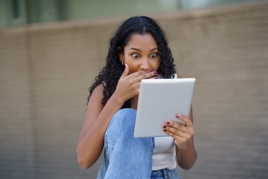 A young woman displays a reaction of surprise while using her tablet, capturing a delightful moment of joy and active engagement in a laidback outdoor setting surrounded by nature