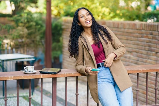 A cheerful young woman with curly hair leans against a railing, holding a phone near a steaming cup of coffee, radiating positivity in a vibrant outdoor setting filled with sunshine