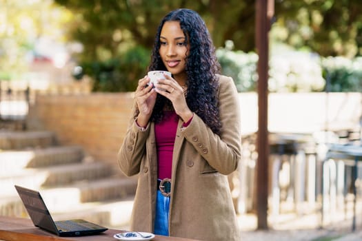 A young woman sits outside, happily sipping a warm cup of coffee as she works intently on her laptop. She embodies a perfect and relaxing worklife balance in a serene and beautiful natural setting