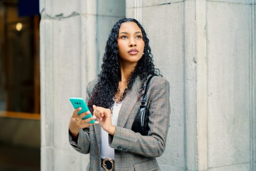 A stylish and fashionable young woman stands confidently against a rustic stone wall while engaging with her smartphone in a dynamic and vibrant city environment, capturing the essence of modern life