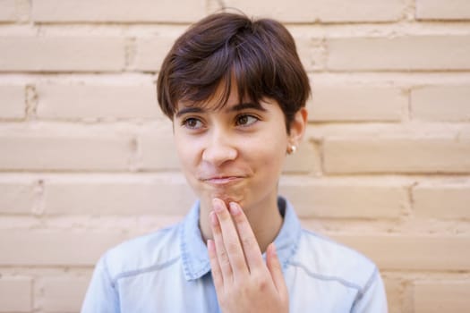 A young individual with stylish short hair, clearly deep in thought, gently rests their hand on their chin, conveying a sense of contemplation while positioned in front of a textured old brick wall