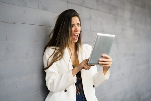 A young woman wearing a stylish white blazer is expressing shock and disbelief while intently looking at her tablet, clearly showcasing a mix of strong emotions against a muted gray background
