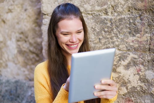 A cheerful young woman in a cozy yellow sweater is happily using her tablet outdoors, smiling and engaged against a charming rustic stone wall, creating a cozy ambiance for relaxation