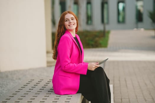 A cheerful and confident businesswoman, dressed in a stylish pink blazer, is sitting on a bench outdoors, happily engaging with her tablet, showcasing professionalism and grace effortlessly