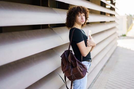 A young woman with beautiful, curly hair is confidently standing against a sleek, modern wall. She holds a phone in one hand and a stylish bag in the other, showcasing a trendy and expressive look