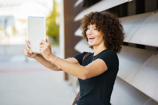 A cheerful, vibrant young woman with lovely curly hair beams brightly while capturing a fun selfie with a tablet, all set against a stylish modern backdrop that enhances her joyful expression