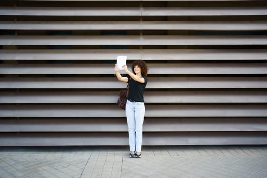 A joyful woman with curly hair holds her smartphone high, ready for a selfie in front of an elegant wall, capturing the vibrant essence of urban life and the pure joy from her radiant smile