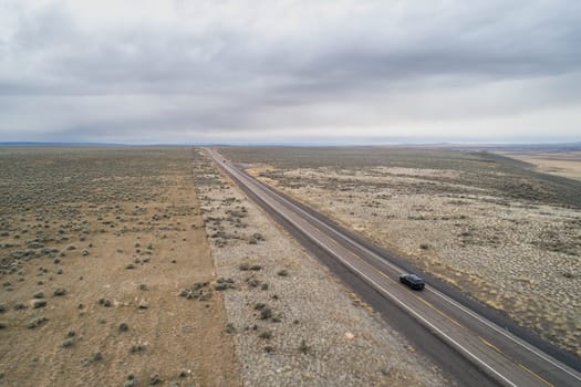 A car is driving down a long, empty road in a desert. The sky is cloudy and the landscape is barren