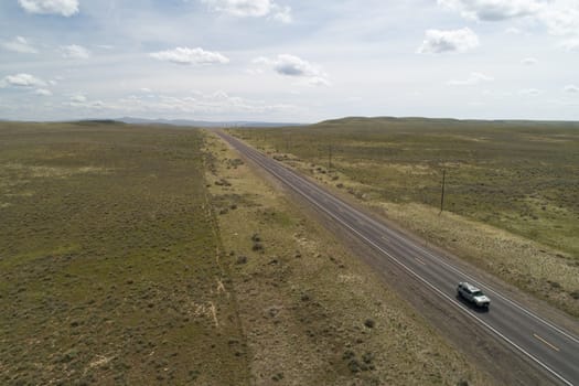A car is driving down a long, empty road in a desert. The sky is cloudy and the landscape is barren