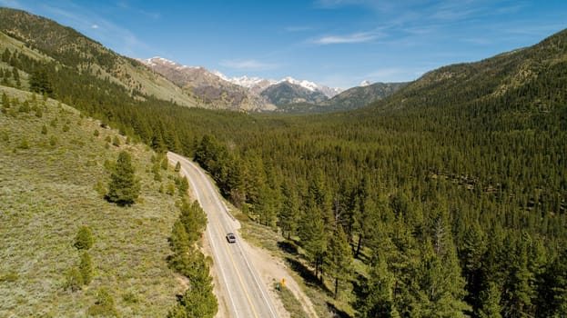 A road with a car driving down it in a forest. The road is surrounded by trees and mountains in the background