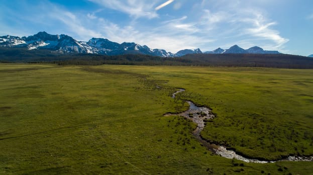 A large, open field with a river running through it. The sky is clear and blue, and the mountains in the background are covered in snow. The scene is peaceful and serene