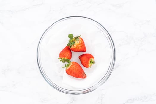Bright red strawberries, interspersed with signs of mold, rest in a glass bowl lined with a paper towel on a white napkin, indicating improper storage techniques.