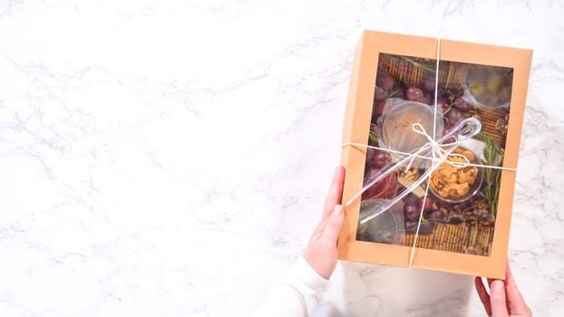 Flat lay. Hands are meticulously adding fresh red grapes to a bowl, complementing a beautifully arranged charcuterie box brimming with a variety of cheeses, olives, and cured meats, set against a sleek marble surface.