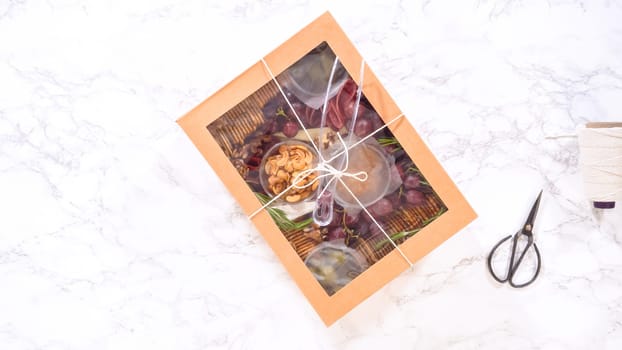 Flat lay. Hands are meticulously adding fresh red grapes to a bowl, complementing a beautifully arranged charcuterie box brimming with a variety of cheeses, olives, and cured meats, set against a sleek marble surface.