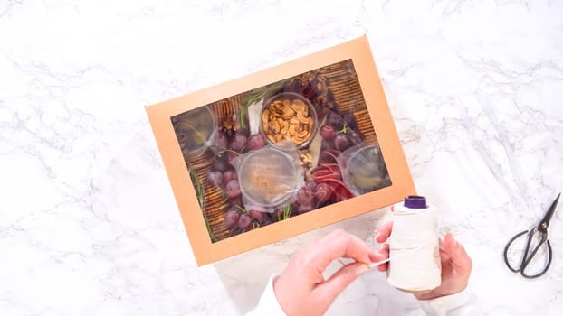 Flat lay. Hands are meticulously adding fresh red grapes to a bowl, complementing a beautifully arranged charcuterie box brimming with a variety of cheeses, olives, and cured meats, set against a sleek marble surface.