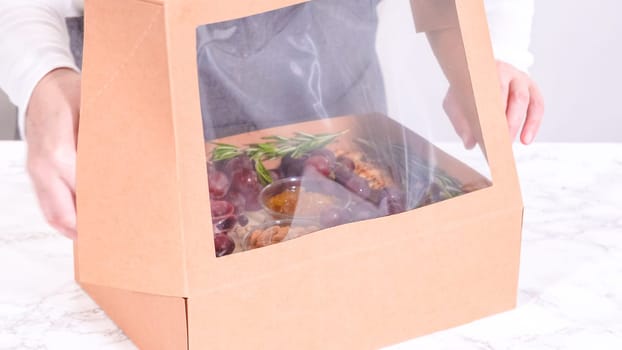 Hands are meticulously adding fresh red grapes to a bowl, complementing a beautifully arranged charcuterie box brimming with a variety of cheeses, olives, and cured meats, set against a sleek marble surface.