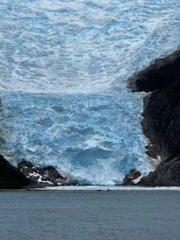 A massive glacier terminus shows textured blue ice as it meets dark ocean water, surrounded by rocky cliffs and melting edges.