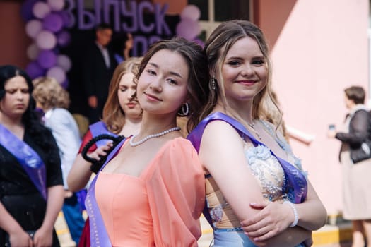Two elegant girls wearing sashes are posing together on their graduation day, celebrating their academic achievement with other classmates