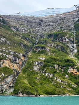 Thin glacial waterfalls stream down rugged, forested cliffs from an icy glacier above, reaching the vivid turquoise waters of a remote fjord below.
