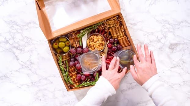 Flat lay. Hands are meticulously adding fresh red grapes to a bowl, complementing a beautifully arranged charcuterie box brimming with a variety of cheeses, olives, and cured meats, set against a sleek marble surface.