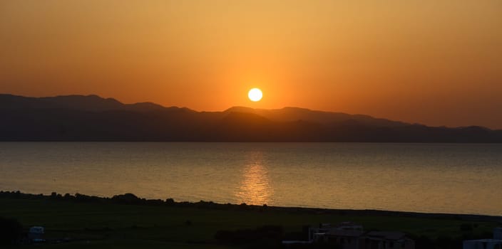 As the sun descends, the sky transforms into warm shades of orange and gold, casting a peaceful glow over calm waters and silhouetted mountains, creating a breathtaking evening view.