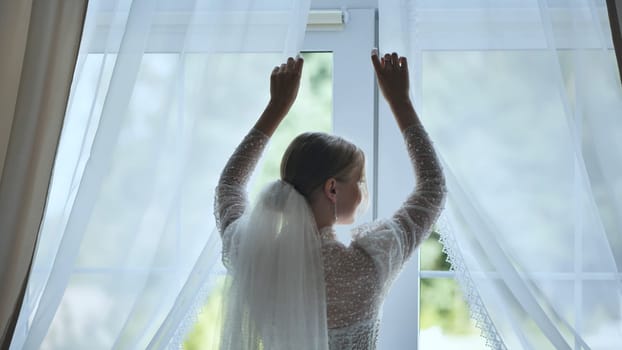 Bride, seen from behind, opens the curtains on her wedding day, revealing a beautiful green garden outside