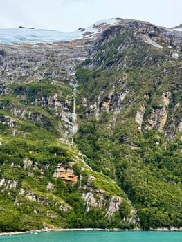 A narrow waterfall flows from a snow-covered glacier down steep rocky cliffs through dense green forest into a bright turquoise fjord below.