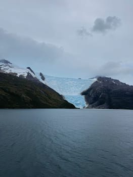 A towering blue glacier stretches from the snow-covered Andes into a tranquil fjord, surrounded by rugged, dramatic Patagonian terrain.