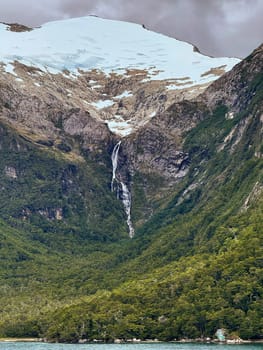 A narrow waterfall cascades from a glacier high in the snowy mountains, cutting through a forested valley and flowing toward a turquoise fjord below.