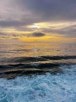 The sun sets over the open ocean, casting golden light on gentle waves and foamy surf below a dramatic sky filled with layered clouds.