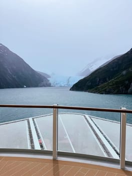 View from a cruise ship deck as it sails through a narrow fjord toward a stunning blue glacier, surrounded by misty mountains and icy waters.
