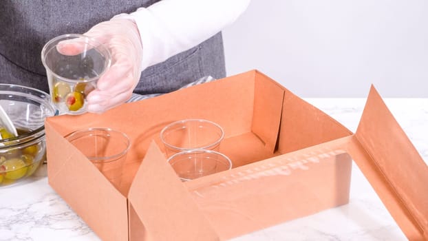 Hands are meticulously adding fresh red grapes to a bowl, complementing a beautifully arranged charcuterie box brimming with a variety of cheeses, olives, and cured meats, set against a sleek marble surface.