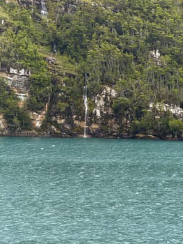 A delicate waterfall flows from a steep, green forested cliff into a vivid turquoise fjord, surrounded by rugged rock and dense vegetation.