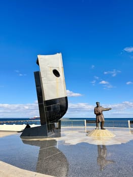 Punta Arenas, Chile 10.22.2023 A modern maritime monument with a bronze sailor statue and ship bow sculpture stands on a reflective plaza by the sea under a clear blue sky.