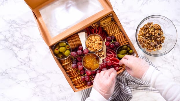 Flat lay. Hands are meticulously adding fresh red grapes to a bowl, complementing a beautifully arranged charcuterie box brimming with a variety of cheeses, olives, and cured meats, set against a sleek marble surface.