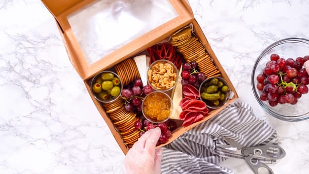 Flat lay. Hands are meticulously adding fresh red grapes to a bowl, complementing a beautifully arranged charcuterie box brimming with a variety of cheeses, olives, and cured meats, set against a sleek marble surface.