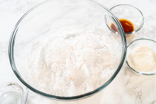 A glass bowl containing powdered sugar sits on a marble countertop, surrounded by small bowls of vanilla extract and other ingredients for preparing Simple White Royal Icing.