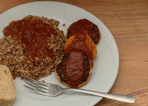 Delightful flavors are presented on a white plate featuring golden patties drizzled with tangy sauce alongside wholesome buckwheat and a slice of fresh bread, perfect for a hearty meal.