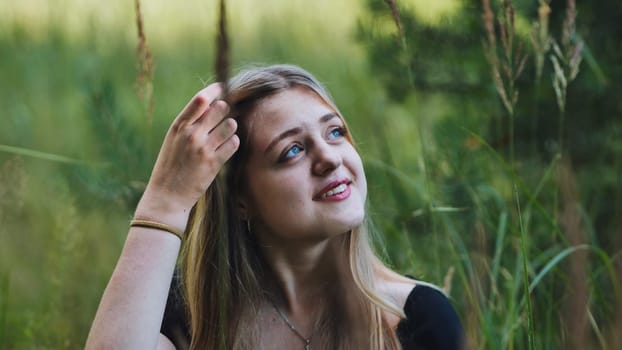 Portrait of a smiling blonde teenage girl touching her hair and looking up, surrounded by tall green grass in a natural outdoor setting