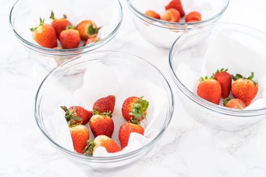 Bright red strawberries, interspersed with signs of mold, rest in a glass bowl lined with a paper towel on a white napkin, indicating improper storage techniques.