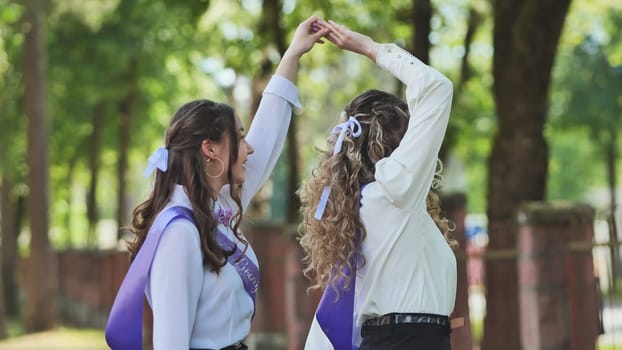 Two happy graduates are holding hands and dancing together, celebrating their last school day in a green park