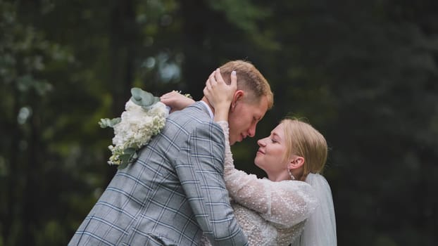 Romantic newlyweds embracing intimately among trees, bride holding delicate wedding bouquet in lush forest setting